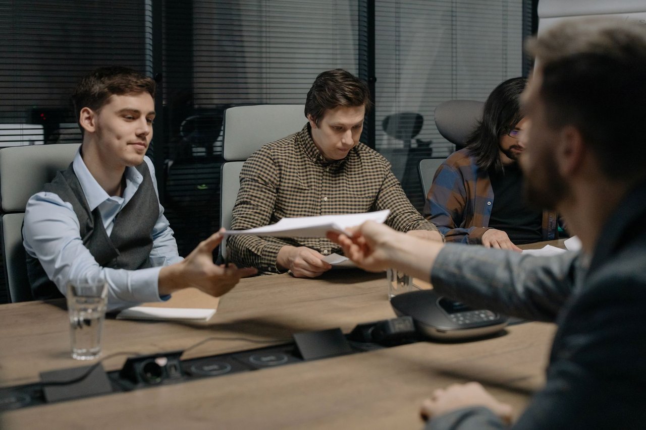 Quatro pessoas trocam documentos durante uma reunião em um escritório.