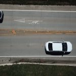 Vista aérea de uma rodovia com dois carros brancos e sinalização direcional nas pistas.