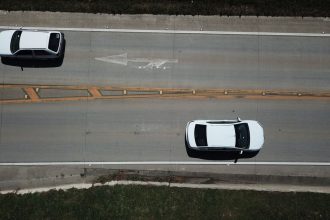 Vista aérea de uma rodovia com dois carros brancos e sinalização direcional nas pistas.